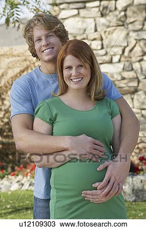 Stock Image - Expectant couple embracing in garden, portrait. Fotosearch