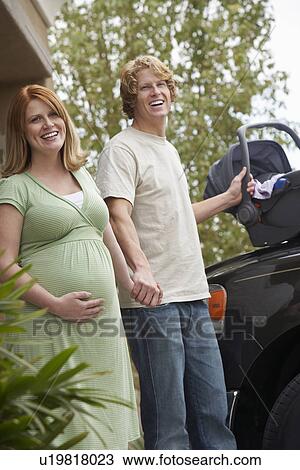 Stock Image - Expectant couple with baby carrier by car, portrait. Fotosearch