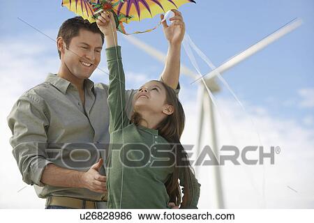 Stock Photograph - Father and daughter (5-6) playing with kite at wind farm. Fotosearch