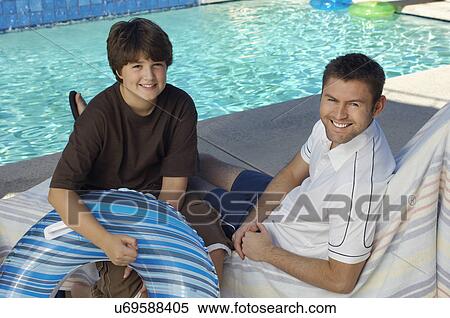 Stock Photography - Father and son holding inflatable raft, sitting by side of swimming pool, portrait. Fotosearch