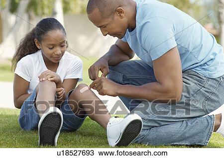 Stock Image - Father applying bandaid to daughter (7-9) in park. Fotosearch