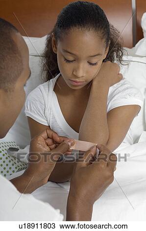 Stock Image - Father applying bandaid to daughter (7-9) in bed. Fotosearch