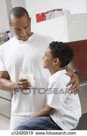 Stock Photo - Father holding glass of milk, embracing son (7-9) indoors. Fotosearch