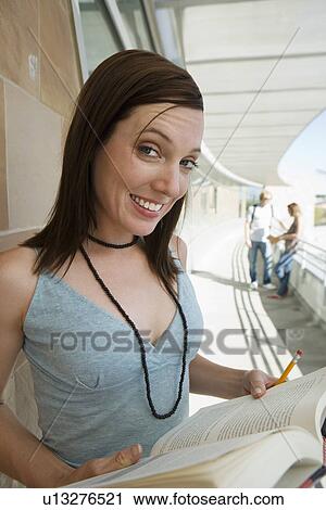 Stock Image - Female student holding book at school, smiling, portrait. Fotosearch