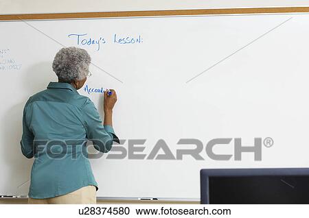 Stock Image - Female teacher writing on whiteboard in classroom. Fotosearch