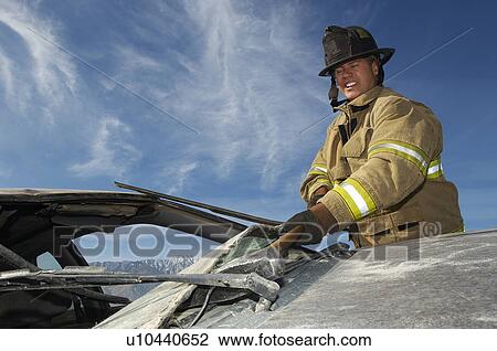 Stock Image - Firefighter hitting crashed car with hammer. Fotosearch