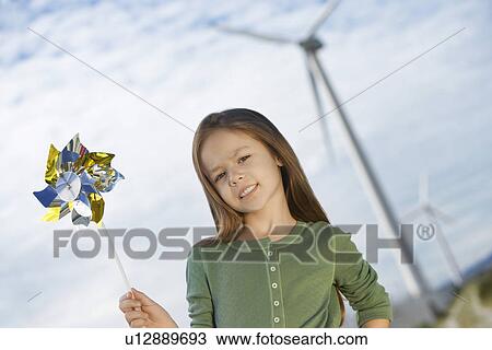 Stock Image - Girl (5-6) holding toy windmill at wind farm, portrait. Fotosearch