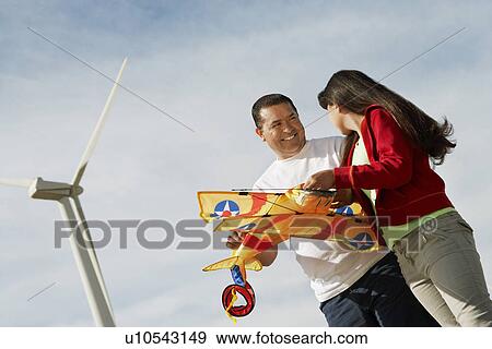 Stock Photo - Girl (7-9) holding airplane kite with father at wind farm. Fotosearch