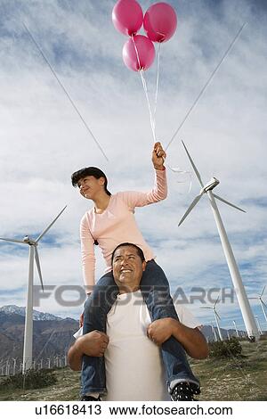 Stock Image - Girl (7-9) holding balloons, sitting on fathers shoulders at wind farm. Fotosearch