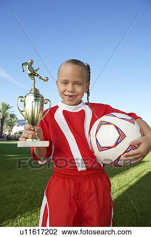 Stock Image - Girl (7-9 years) soccer player holding trophy and ball, portrait. Fotosearch