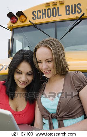 Stock Image - High School Girls Looking at Laptop by School Bus. Fotosearch