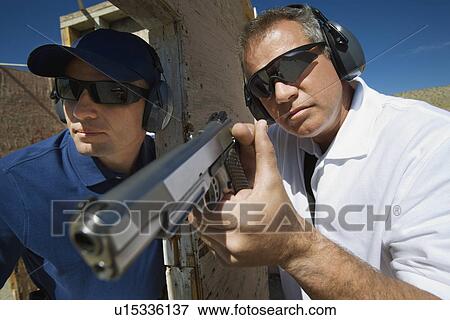 Stock Photo - Instructor assisting man aiming hand gun at firing range. Fotosearch