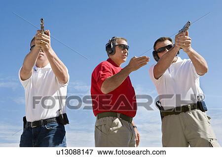 Stock Photo - Instructor assisting men aiming hand guns at firing range. Fotosearch