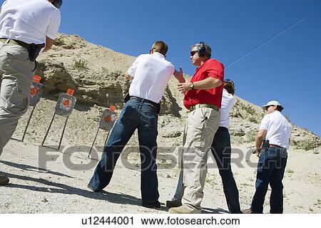 Stock Image - Instructor assisting people aiming guns at firing range. Fotosearch
