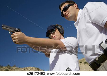 Stock Image - Instructor assisting woman aiming hand gun at firing range, low angle view. Fotosearch