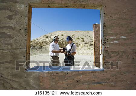 Stock Image - Instructor assisting woman loading gun at firing range in desert. Fotosearch