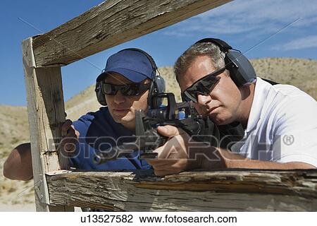 Stock Image - Instructor with man aiming machine gun at firing range. Fotosearch
