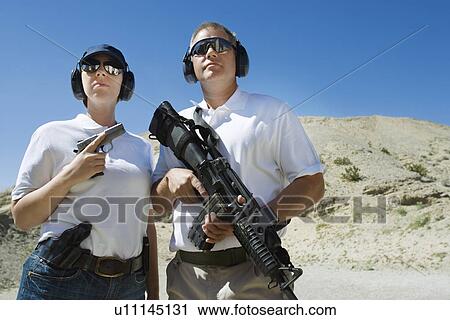 Stock Image - Man and woman holding guns at firing range in desert. Fotosearch