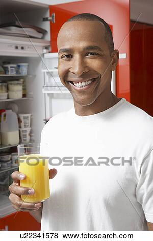 Stock Photo - Man holding glass of orange juice, by fridge, portrait. Fotosearch