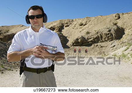 Stock Image - Man holding hand gun at firing range, portrait. Fotosearch