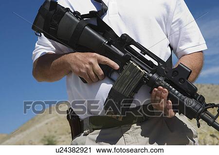 Stock Image - Man holding machine gun at firing range, mid section. Fotosearch
