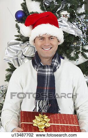 Man in Santa hat holding present in front of Christmas tree View Large Photo Image Stock Image - Man in Santa hat holding present in front of Christmas tree. Fotosearch