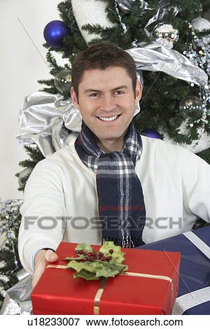 Man offering present in front of Christmas tree View Large Photo Image Stock Photo - Man offering present in front of Christmas tree. Fotosearch