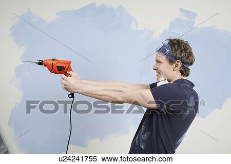 Stock Photography - Man pointing drill in front of half painted wall, side view. Fotosearch