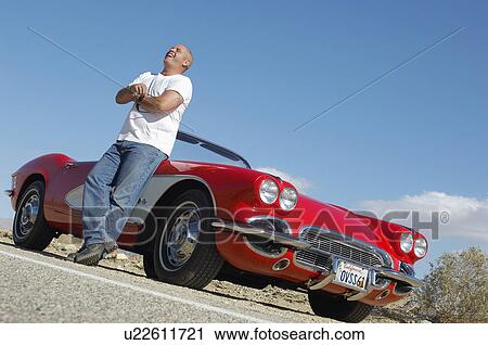 Stock Image - Man standing beside classic car on road. Fotosearch