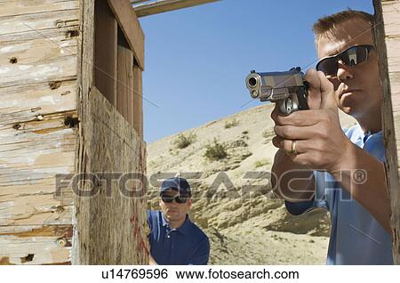 Stock Photograph - Man watching colleague aiming hand gun at firing range. Fotosearch