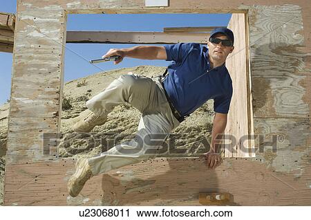 Stock Image - Man with hand gun jumping obstacle at firing range. Fotosearch