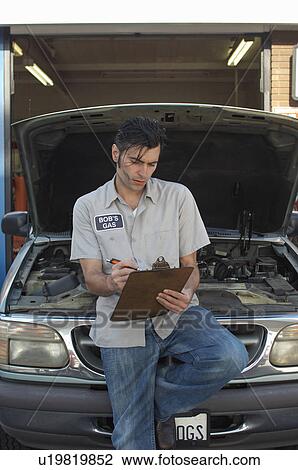 Stock Image - Mechanic writing on clipboard by open car hood. Fotosearch