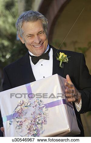 Stock Photo - Middle-aged man in tuxedo holding present, portrait. Fotosearch