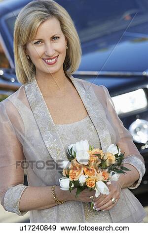 Stock Photo - Middle-aged woman holding bouquet in front of limousine, portrait. Fotosearch