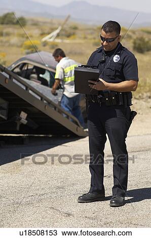 Stock Image - Police officer writing notes, tow truck driver lifting crashed car in background. Fotosearch