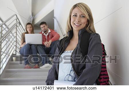 Stock Image - Portrait of female student on stairs, friends using laptop in background. Fotosearch