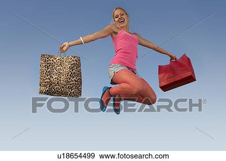 Stock Photo - Portrait of young woman jumping with shopping bags in park. Fotosearch