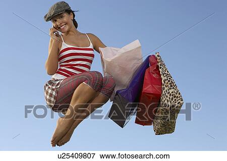Stock Photo - Portrait of young woman jumping with shopping bags, using mobile. Fotosearch