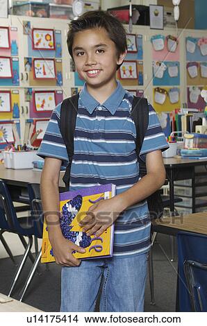 Picture - School boy holding book in classroom, portrait. Fotosearch