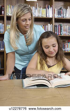 Stock Photograph - School girl reading book with teacher in library. Fotosearch