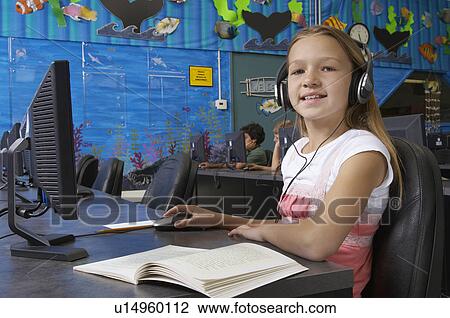 Stock Image - School girl wearing headphones in computer room, portrait. Fotosearch