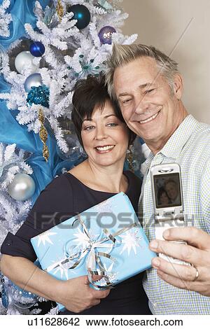 Stock Image - Senior couple taking picture with cell phone in front of Christmas tree. Fotosearch
