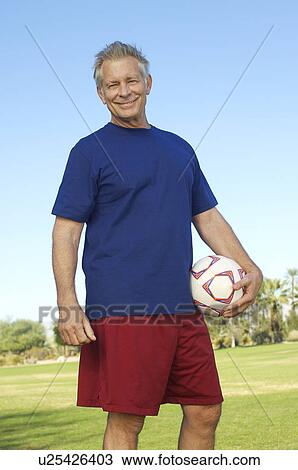 Stock Image - Senior man holding soccer ball in park, portrait. Fotosearch