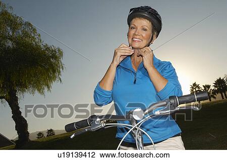Stock Image - Senior woman adjusting cycling helmet at dusk, portrait. Fotosearch