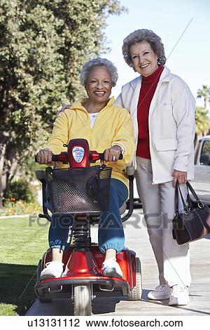 Stock Image - Senior woman on motor scooter on urban street with friend. Fotosearch