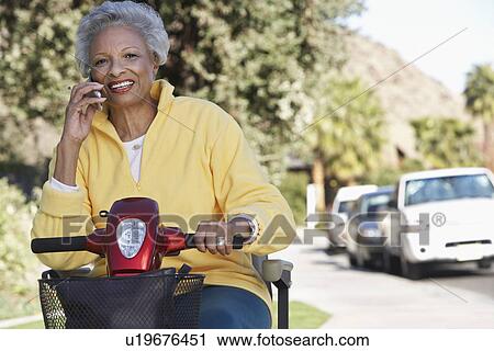 Stock Image - Senior woman on motor scooter on urban street, talking on mobile phone. Fotosearch