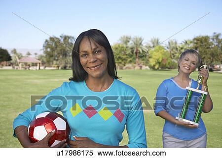 Stock Photography - Senior women holding soccer ball and trophy in park, portrait. Fotosearch