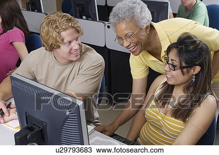 Stock Image - Teacher helping two students working in computer classroom. Fotosearch