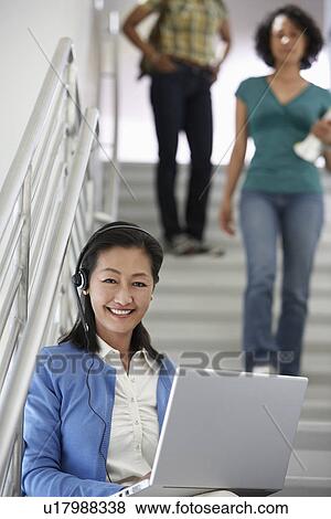 Stock Photo - Teacher using laptop on stairs in school, students in background. Fotosearch