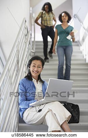 Stock Photography - Teacher using laptop on stairs in school, students in background. Fotosearch